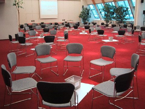 Das Bild zeigt einen großen, modernen Workshopraum, indem Arbeitsgruppen in Sesselkreisen für eine Klausur vorbereitet sind. Der Teppich ist rot.
